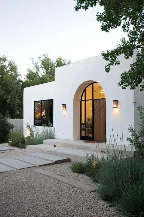 Modern spanish house in white stucco modern arched windows in black trim large handcrafted wooden front door modern lamp fixtures gravel yard with. Unveil the charm and elegance of Spanish houses, and find out how their timeless beauty can inspire your home design. Arch Garage Doors House, Desert Houses Architecture, Stucco Exterior Before And After, Transitional Architecture Exterior, Modern Hacienda Exterior, Exterior Arched Windows, Modern Spanish House Interior, Modern Front House Design, Mexican Exterior Houses