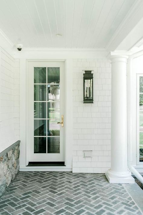 Covered patio boasts gray brick herringbone pavers located beneath a white plank ceiling help up with Doric columns. White Brick Walkway, Front Porch With Pavers, Gray Brick Patio, White Brick Porch, Herringbone Paver Patio Ideas, Herringbone Pavers Walkway, Grey Brick Patio, Herringbone Pavers, White Brick Patio