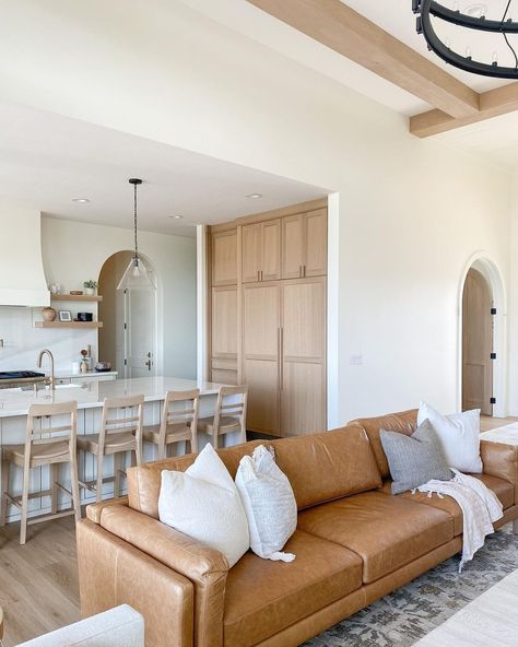 High white ceilings with light wood beams frame this living room and kitchen. A brown leather couch accessorized with white, beige, and grey throw pillows rests on a patterned rug. Behind the sofa is a large island with glass pendant lights and light, wood cabinets along one wall. Light Brown Leather Couch, Tan Leather Couch Living Room, Tan Couch Living Room, Leather Sectional Living Room, Brown Leather Couch Living Room, Leather Couches Living Room, Brown Couch Living Room, Beach Living Room, Leather Sofa Living Room