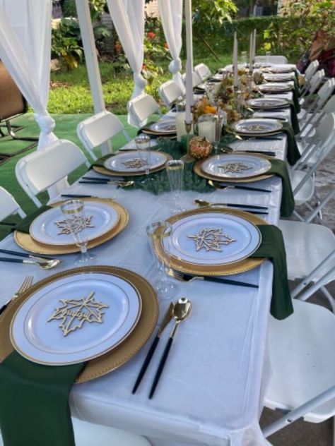 Exude a surreal appeal at your outdoor party with a sophisticated & elegant tablescape. Drape the table with a white tablecloth & stylishly set a moss-green cheesecloth runner in the middle. Also, arrange gorgeous blooms to impart an ethereal appeal & inset taper candles in gold candlestick holders to radiate a subtle glow. Elevate the place setting with gold chargers, gold rim white plates & clear flutes, gold cutlery with black handles, & moss green napkins for graceful tabletop decor. Green And White Thanksgiving Table, White Green And Gold Table Setting, Green Cream And Gold Wedding Table Settings, Green And Gold Table Setting, Green And Gold Wedding Place Settings, Green And Gold Place Setting, Gold Charger Green Napkin, Gold Table Decor, Green Table Settings