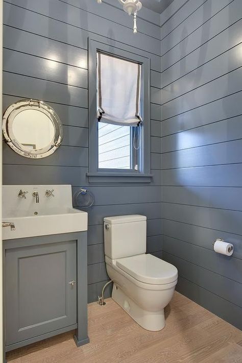 Blue cottage powder room boasts blond oak wood floors framed by blue shiplap walls and is fitted with a white porcelain toilet positioned beneath a window dressed in a gray grosgrain roman shade. Shiplap Powder Room, Cottage Powder Room, Blue Shiplap, Shiplap Bathroom Wall, Blue Walls Living Room, Vertical Shiplap, Farmhouse Bathroom Decor Ideas, Shiplap Bathroom, Shiplap Walls