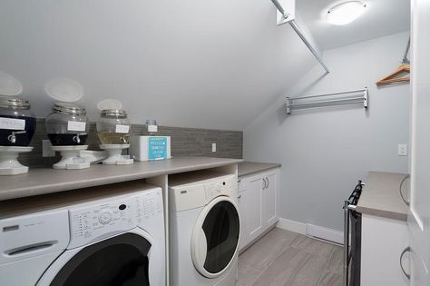 An attic laundry room features white cabinetry paired with gray countertops and a gray glass tiled backsplash placed under a sloped ceiling. Gray Countertops, Farmhouse Laundry Room Ideas, Attic Wardrobe, Glass Closet Doors, Attic Renovation Ideas, Glass Closet, Attic Closet, Attic Playroom, Attic House