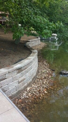 We tore out an old wooden reatining wall and replaced with a tumbled retaining wall block. We placed creek rock on the front side of the wall for a visual apperance and to also hide the concrete footing. We also extended the paver walkway, attached the dock, and added topsoil to freshen everything up. Located in central MO.  ** www.greenacresmidmo.com *** Creek Retaining Wall, Pond Retaining Wall Ideas, Lake Retaining Wall Ideas, Seawall Ideas, Lake Landscaping, Lake Dock, Paver Walkway, Landscaping Retaining Walls, Hillside Landscaping