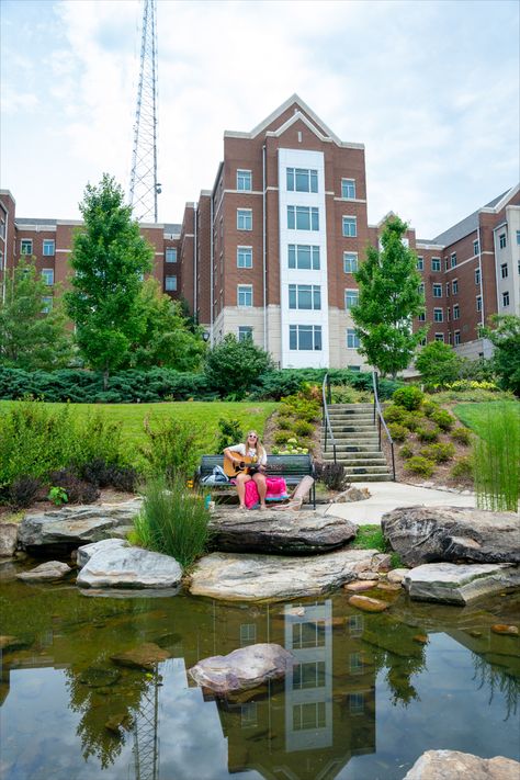 Belmont University Bear creek; pond with stones and muddy water. Female student sitting on a bench playing the acoustic guitar. Large dorm building in the background with blue sky and telephone pole. Belmont University Nashville, Nashville Aesthetic, Belmont University, College Tour, Visit Nashville, Spring Semester, Nashville Trip, College Town, Spring Photos