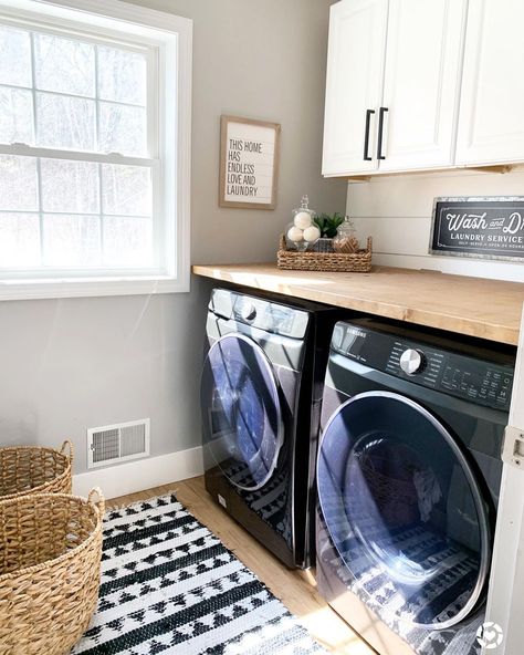 Black washer and dryer are positioned underneath a light wood laundry countertop against a white shiplap wall. White cabinets with black pulls are mounted above the counter in this laundry room with a black and white patterned rug. Black Washer And Dryer, Laundry Countertop, Folding Station, Laundry Room Counter, Laundry Room Folding Table, Laundry Room Countertop, Laundy Room, Laundry Makeover, White Laundry Rooms