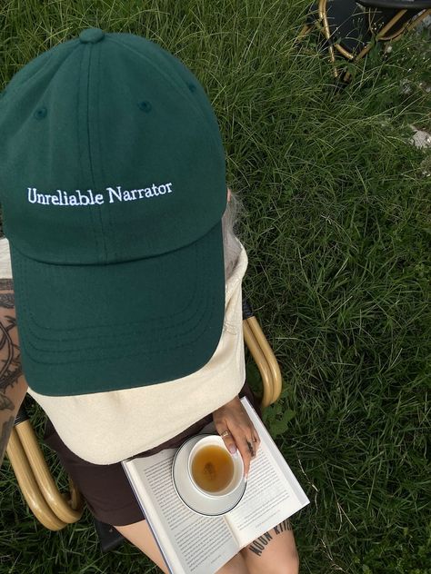 Overhead photo of a green hat that reads 'Unreliable Narrator' worn by a girl with a book and a cup of tea in her lap. She's sitting in a chair in a field. Unreliable Narrator, Soft Spot, Good And Bad, Bad Hair Day, Bad Hair, Baseball Hat, Dad Hat, Hair Day, Trucker Cap