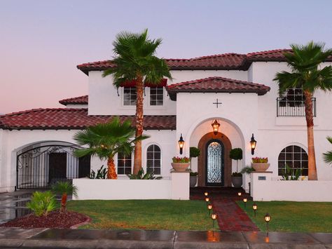 The exterior of this traditional Spanish-style home has a brick walkway, an arched entryway and a red tile roof. Landscape lighting illuminates the entry to the home and iron gate details can be seen on the left. Exterior Home Lighting, Spanish Style Home Exterior, Modern Spanish Style Homes, Mediterranean Revival Architecture, Mediterranean Exterior Homes, Mediterranean House Designs, Mediterranean Homes Exterior, Mediterranean Exterior, Best Exterior Paint