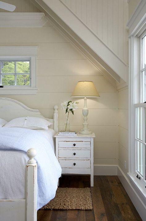 Beautiful monochromatic bedroom with white distressed bed and white distressed nightstand. The bed is layered with ivory bedding topped with a folded white duvet. The distressed nightstand is topped with a tall ivory table lamp, stacked books and a vase of white lilies. The walls are paneled with wood and painted a rich buttermilk color with white trim. A small window adorns the wall over the headboard. The hardwood floors are layered with a small braided sisal rug. Coastal Bedrooms, Cottage Bedroom, Coastal Bedroom, White Cottage, Cottage Interiors, White Bedroom, Beautiful Bedrooms, Cottage Homes, Cheap Home Decor