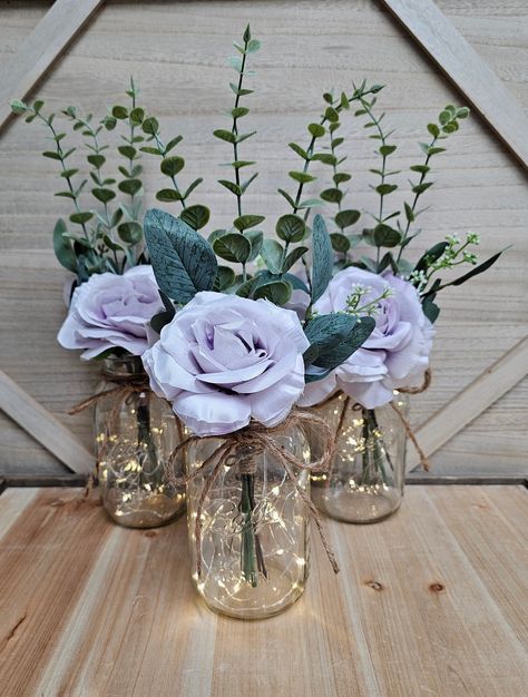 This beautiful 3 piece set of faux rose and eucalyptus quart size mason jar centerpieces are a Glassy Gal original design. Each centerpiece includes one set of warm white LED fairy lights, a jute bow, a deluxe faux rose and eucalyptus greenery arrangement.  These centerpieces are very popular for weddings, baby showers, parties, corporate events, holiday events, and any other event that requires table centerpieces. They also look great in any room of your house or office. To see other lighted ja Purple Flower Candle Centerpieces, Light Purple Centerpiece Wedding, Wedding Decorations Purple And White, Purple And Grey Centerpieces Wedding, Lavender And Light Green Wedding, Lavender Themed Bridal Shower Ideas, Lilac And Sage Wedding Centerpieces, Purple Baby Shower Centerpieces, Mauve Centerpieces