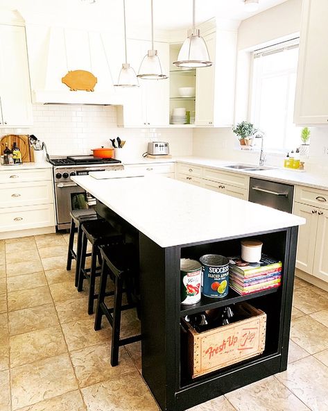 A kitchen with tan floor tiles and white cabinets features a black contrasting kitchen island with open shelving on the end panel. Nickel and glass pendant lights hang over the island's white countertop with black backless stools tucked underneath. Cabinet End Panel Ideas, Island With Open Shelving, Island End Panel, Kitchen Island End Panels, Island Shelf, Kitchen Island Ends, Contrasting Kitchen, Contrasting Kitchen Island, Kitchen Island Layout