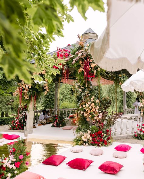 Romantic outdoor Sikh wedding ceremony with a gazebo adorned with lush greenery, cascading red and pink florals, soft drapery, and vibrant pink cushions lining a gold reflective aisle, creating a stunning and intimate atmosphere. Outdoor Sikh Wedding, Floral Gazebo, Wedding Ceremony Setup, Serene Garden, Red Peach, Romantic Atmosphere, Sikh Wedding, Red Pillows, Garden Set