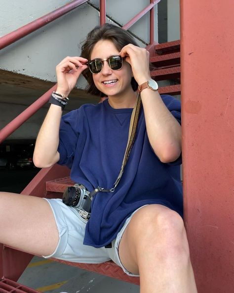 Mak Ingemi is sitting on some red stairs. She is wearing a blue shirt, cream shorts and sunglasses. She is looking and smiling towards the camera. She has her fingers on her sunglasses, as if she wants to take them off. Mak Ingemi Outfit, Lesbian Swim Outfit, Soft Masc Lesbian Fashion, Lesbian Short Hair, Masc Lesbian Fashion, Masc Outfits For Women, Soft Masc, Swimming Outfits, Masc Women