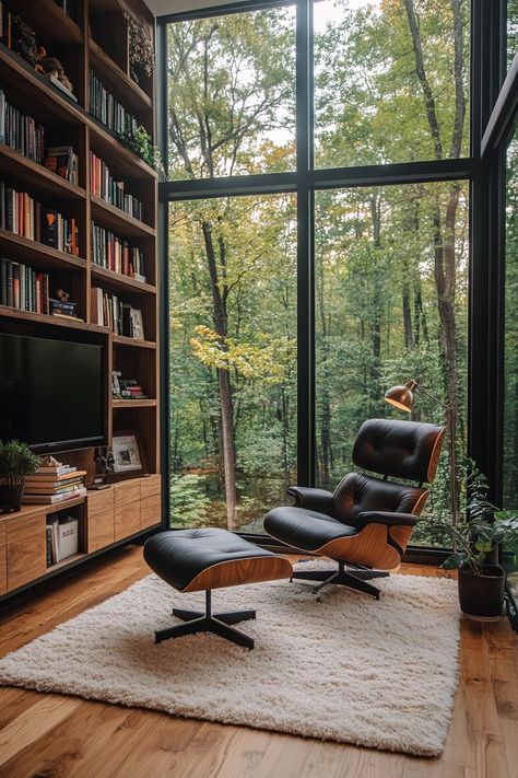 A modern home library with wood and black steel furniture, a white rug, built-in bookshelves, a modern TV, large windows offering a forest view, an Eames chair, a reading light, and a minimalist design. Room With Forest View, Black And Wood House Interior Design, Eames Chair Living Room, Wood And Steel Furniture, Apartment Library, Beautiful Estates, Modern Home Library, Library Modern, Library Home