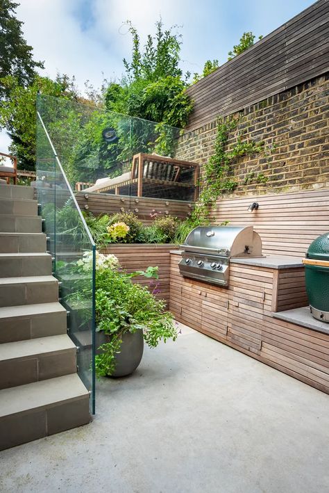 This outdoor kitchen situated in the basement area of a Clapham garden exudes a clean and stark, yet warm and inviting charm. Surrounded by vibrant greenery and bathed in the soft glow of dappled sunlight, this area blends the comforts of an indoor kitchen with the refreshing allure of the great outdoors. It invites you up the stairs to the garden above. Steep Gardens, Townhouse Garden, Sloped Backyard, Tiered Garden, Garden Levels, Minimalist Garden, Back Garden Design, Sloped Garden, London Garden