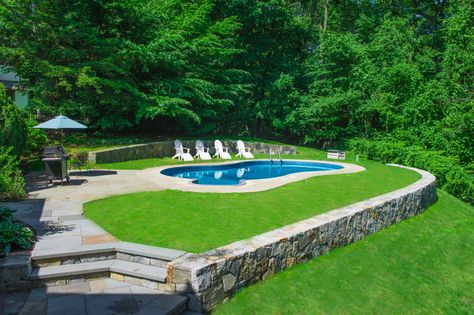 A pool surrounded by beautiful green lawn Sloped Pool, Retaining Wall Around Pool, Pool Retaining Wall, Deck Addition, Deck Landscaping, Inground Pool Landscaping, Sloped Yard, Sloped Backyard, Stone Retaining Wall