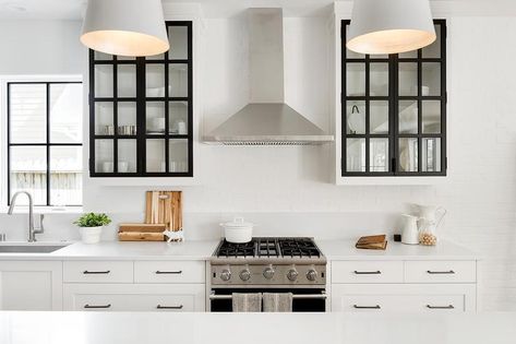 Black and white glass frame kitchen cabinets display white tableware flanking a stainless steel vent hood above a range, white shaker cabinets, and white quartz countertops. Black Metal And Glass Kitchen Cabinets, Metal Glass Kitchen Cabinets, Black Glass Upper Cabinets, Kitchen Cabinet Display, Black Glass Cabinet, Brown Kitchen Designs, Framed Kitchen Cabinets, Metal Kitchen Cabinets, Glass Kitchen Cabinet Doors