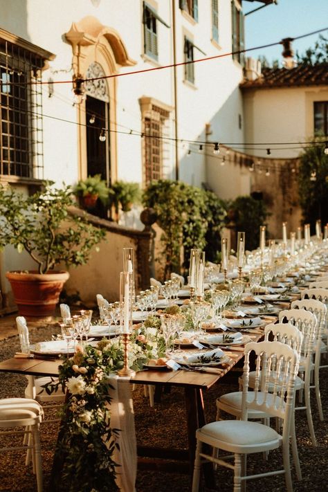 Wedding reception with a beautiful family style long wooden table, white romantic chairs, table runner made of eucalyptus, tea roses and white garden flowers, white taper candles in golden candelholders and vintage table settings under the string lights at a beautiful Italian garden of the 17th century Tuscan villa. White Chairs Wedding, Vintage Table Settings, White Garden Flowers, White Taper Candles, Family Style Weddings, White Flowers Garden, Family Style Table, Long Table Wedding, Vintage Table Setting