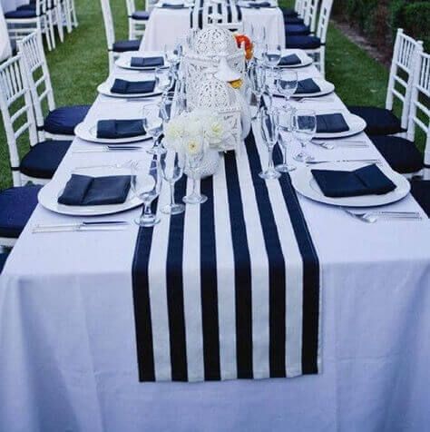 Glorify your outdoor party tables with an imperial demeanor using white rectangular tablecloths coupled with striking white and navy blue striped satin runners. Accentuate this table setup by placing white acrylic charger plates paired with navy blue linen napkins, wine glasses, and silver cutlery. Also, place white Moroccan lanterns on the table and bedeck the white armless chairs with navy blue padded seat cushions to elevate the event table setup to the next level. Striped Tablecloth, Outdoor Bridal Showers, Nautical Bridal Showers, Preppy Wedding, Fiesta Bridal Shower, Aisle Runners, Striped Tablecloths, Bridal Shower Tables, Wedding Tablecloths