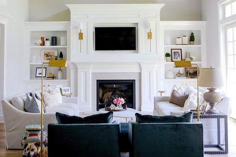 Light gray slipcovered sofas sit facing an oval white and gold coffee table placed in front of a white fireplace mantel fixed beneath a TV niche flanked by brass sconces. Fireplace Sconces, White Fireplace Mantels, Elegant Family Room, White Built Ins, Built In Around Fireplace, Navy Sofa, Fireplace Bookshelves, Built In Shelves Living Room, Marble Fireplace Surround