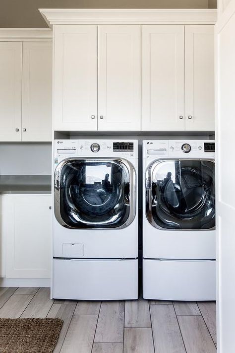 A white front loading washer and dryer is placed on wood like floor tiles and enclosed beneath white shaker cabinets accented with small polished nickel square knobs and lined with white molding. Cabinets Over Washer And Dryer, Raised Washer And Dryer Ideas, Washer And Dryer Pedestal, Blue Laundry Rooms, Laundry Room Paint Color, Mudroom Remodel, White Molding, Laundry Cabinets, Laundry Room Layouts