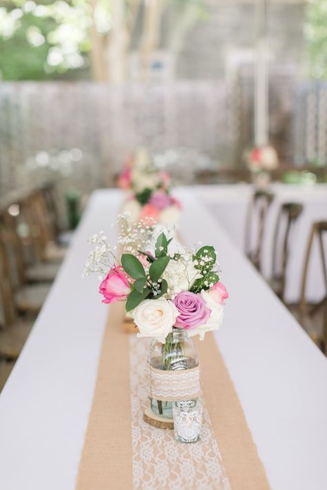 Burlap and lace table runners sitting int he center of white table clothes clear glass mason jar vases sitting on top of wood disks. Inside of the jars are White roses, pink roses, orange roses, and purple roses. Wedding reception, table centerpieces. Mason Jar Roses Centerpieces, Easy Floral Centerpieces, Wedding Mason Jar Ideas, Mason Jar Flower Centerpieces, Mason Jar Wedding Centerpieces, Engagement Party Centerpieces, Purple Roses Wedding, Mason Jar Center, Mason Jar With Flowers