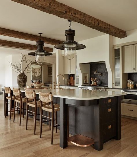 Kitchen features vintage bronze lanterns that illuminate a honed marble top oval island with antique brass deck mount gooseneck faucet and abaca counter stools, a black and white cooking alcove, white cabinets with bronze pulls and wooden ceiling beams. Oval Island Kitchen, Oval Kitchen Island, Cooking Alcove, Island Nook, Oval Island, Wooden Ceiling Beams, Kitchen Island Finishes, Tan Kitchen, Wooden Island