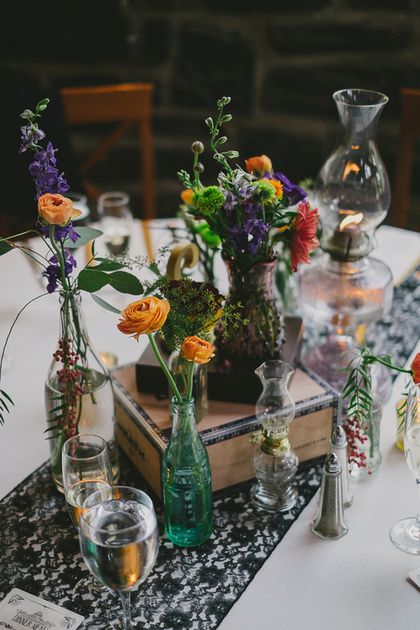 Bold wedding centerpieces with black lace table runner. Antique oil lamps and bottles filled with flowers and stacked cigar boxes with gold and brass accents. Ranunculus, roses, eucalyptus. Wedding flowers in plum, burgundy, red, yellow and green.  Floral Design: www.papertini.com  Photography: www.adamchapinphotography.com Black Table Runners, Oil Lamp Centerpiece, Gatsby Wedding Decorations, Country Wedding Centerpieces, Purple Wedding Centerpieces, Vintage Wedding Centerpieces, Lace Ideas, Purple Bridal Shower, Bottle Centerpieces