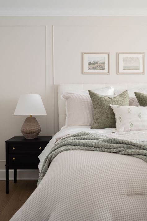 A serene bedroom featuring a neatly made bed with soft, textured white bedding and green accent pillows. A cozy green throw is draped over the bed, adding a touch of nature-inspired color. A black nightstand with gold hardware holds a neutral-toned lamp. Above the bed, two framed landscape artworks hang on the light cream-colored wall, contributing to the peaceful and minimalist aesthetic of the room. The overall atmosphere is calm and inviting, perfect for relaxation. Bedroom With Sage Green Accents, Sage Green Accents, Minimalistic Decor, Natural Vibes, Serene Bedroom, Bedroom Retreat, Of Montreal, English House, Cozy Bedding