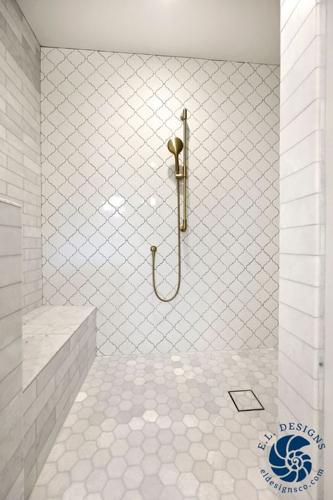 The image looks into a white open shower space. There is subway tile on the walls with an accent wall of glazed porcelain arabesque tile straight ahead. The shower floor is a marble hexagon tile, and the drain is a tile-in drain. The plumbing fixtures are brass and features a sitting bench on the left. White Subway Tile Shower With Marble Hexagon Floor, Arabesque Tile Shower Wall, Arabesque Tile Shower, Arabesque Tile Bathroom, Hexagon Bathroom, Arabesque Tiles, White Subway Tile Shower, Marble Subway Tile, Marble Porcelain Tile