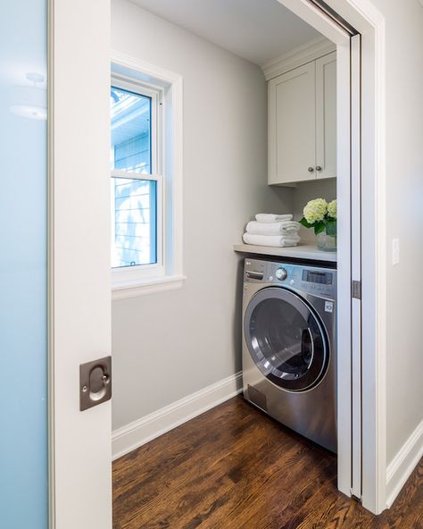 Pocket doors open to small mudroom boasting gray walls framing a window flanked by face to face washer and dryer situated under gray countertop doubling as folding station under shaker cabinets atop oak stained wood floors. Laundry Nook, Laundry Room Storage Shelves, Tiny Laundry Rooms, Laundry Room Doors, Grey Countertops, Laundry Room Cabinets, Laundry Closet, Small Laundry Rooms, Small Laundry