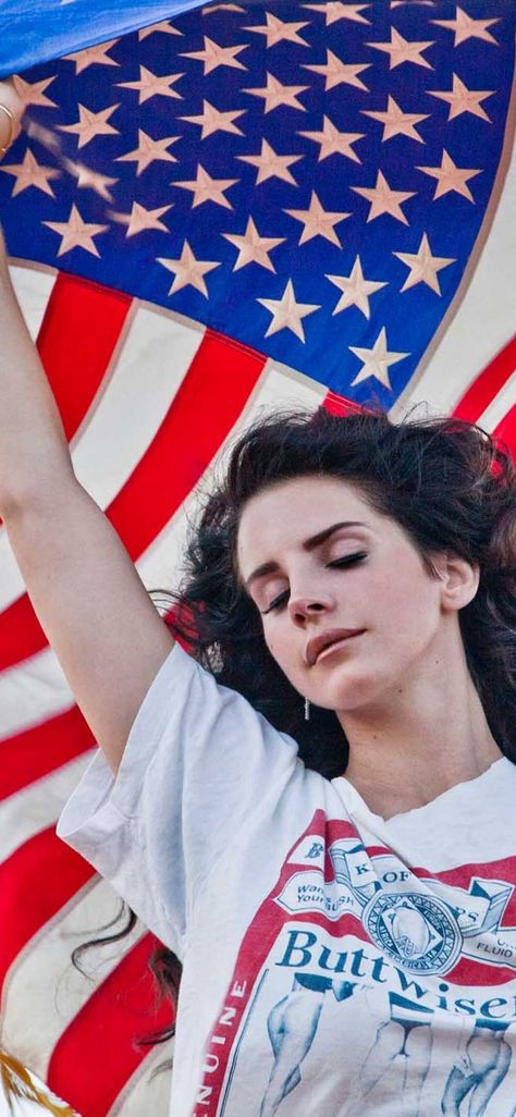 A white brunette, singer/songwriter Lana Del Rey,  stands with her eyes closed and wind blowing through her hair. She is holding the American flag above herself, letting it flow in the wind, and wearing a shirt that says Buttwiser, a spoof of Budweiser beer. Ride Lana Del Rey, Lana Del Rey Ride, Lana Del Rey Quotes, Iphone Wallpaper Music, Lana Del Rey Love, Lana Del Rey Lyrics, Man Wallpaper, Vintage Americana, Lana Del Ray