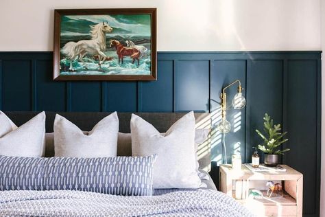 A dark wood-framed art piece is mounted above this gray upholstered bed clad in white and blue bedding. The bed is accompanied by a light wood bedside table lit by a gold and glass wall sconce. Add depth to the room with navy blue board and batten paneling behind the bed. Blue Board And Batten, White And Blue Bedding, Beige Living Room Walls, Gray Upholstered Bed, Light Wood Bed, Grey And White Bedding, Colorful Bedroom, White Wainscoting, Light Blue Walls