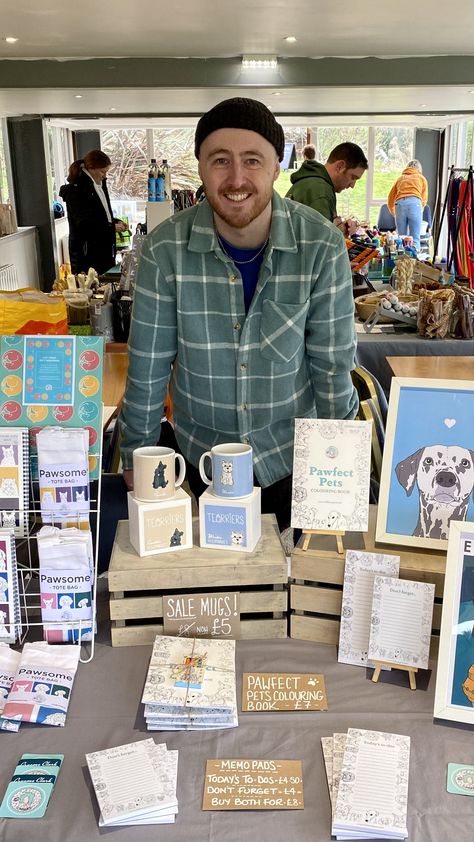 The photo is of me, Graeme Clark aka Oldmangrey Illustration, standing behind a market stall at an indoor event (though I still have my beanie hat on as it was cold!) The table has a grey tablecloth and the section that can be seen has colouring books, memo pads, framed pet prints, folded tote bags, mugs, and business cards, plus some price point signage. Farmers Market Dog Display, Dog Market Stall, Animal Stall Decoration Ideas Fair, Flea Market Stall Ideas, Pet Exhibition Booth Design, Miniature Market Stalls, Art Stall, Dog Marketing, Stall Display