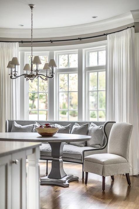 White curtains cover windows framed by a gray wall and positioned behind a curved dining settee topped with gray pillows and positioned beside a light gray skirted dining chair facing a white and gray oval pedestal dining table. Bay Window Curtain Ideas, Curved Banquette Seating, Bay Window Dining, Dining Room Bay Window, Curved Banquette, Bow Windows, Bay Window Treatments, Bay Window Ideas, Settee Dining