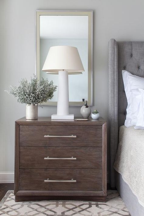 Restful bedroom features a brown 3 drawer nightstand placed on a gray trellis rug and fitted with satin nickel pulls lit by a white lamp positioned in front of a gold and gray mirror mounted to a light gray wall beside a gray linen tufted wingback headboard and bed dressed in cream bedding. Bedroom Gray Headboard, Gray Headboard, Gray Bedroom Walls, Brown Nightstands, Restful Bedrooms, Bedroom Gray, Grey Headboard, Transitional Bedroom, Trendy Diy