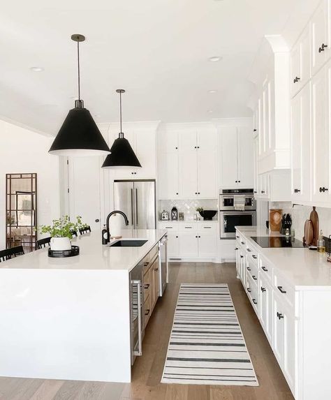 Black knobs and pulls adorn white kitchen cabinets with white countertops. The cabinets frame a white island lit by black pendant lights in this L-shaped kitchen with glazed white herringbone tile backsplash. White Herringbone Tile Backsplash, Extra Space Storage, Black Cabinet Hardware, Black Kitchen Cabinets, Space Storage, White Countertops, Kitchen Inspiration Design, White Kitchen Cabinets, Farmhouse Style House