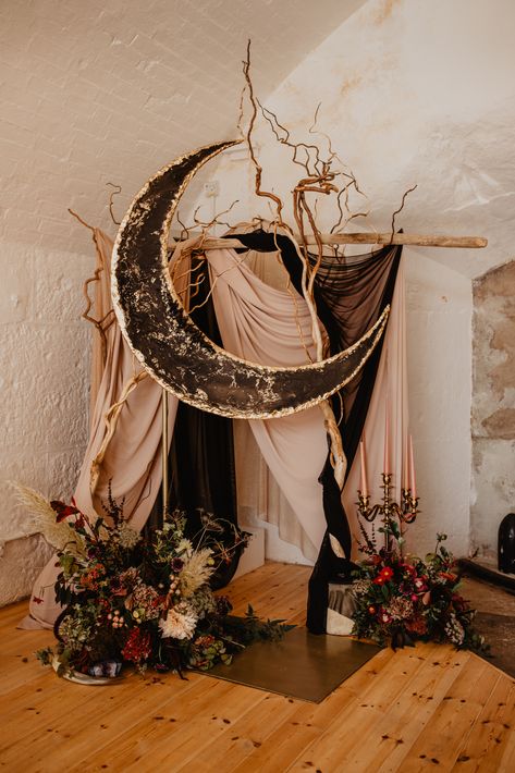 A large crescent moon in black and gold resin, in a geode style sits high in front of winding branches and drapes. Deep florals sit at the base of the ceremony arch, with a large golden candelabra with 5 candle sticks. Winter Solstice Wedding, Celestial Backdrop, Solstice Wedding, Celestial Wedding Theme, Starry Night Wedding, Festival Bride, Moon Wedding, Celestial Wedding, Goth Wedding