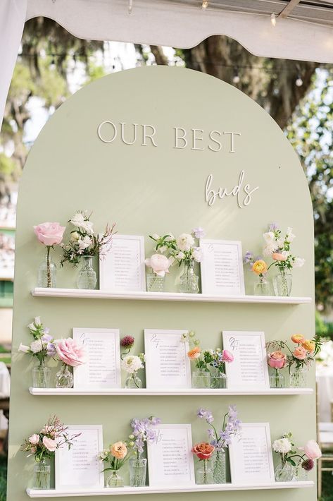 Romantic wedding seating chart display with a soft green backdrop, pastel floral arrangements, and framed guest lists for a modern yet timeless touch. Perfect for outdoor, garden, or elegant wedding designs, this creative idea combines stylish functionality and stunning decor. Seat Wedding Chart, Bookcase Wedding Seating Chart, Seating Chart Ideas For Small Wedding, Wildflower Seating Chart Wedding, Pastel Wedding Seating Chart, Spring Seating Chart, Table List Wedding Seating Charts, Seating Chart Garden Wedding, Floral Wedding Seating Chart