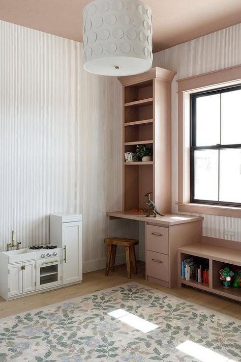 Kid's room features a pink built in window seat next to a built in desk under shelves and a white kitchenette with a floral rug lit by a white pendant hung from a pink ceiling. White Kitchenette, Desk Under Window, White Family Rooms, Flat Front Cabinets, Built In Desk And Shelves, Pink Girls Room, Blue Stripe Wallpaper, Built In Window Seat, Pink Ceiling