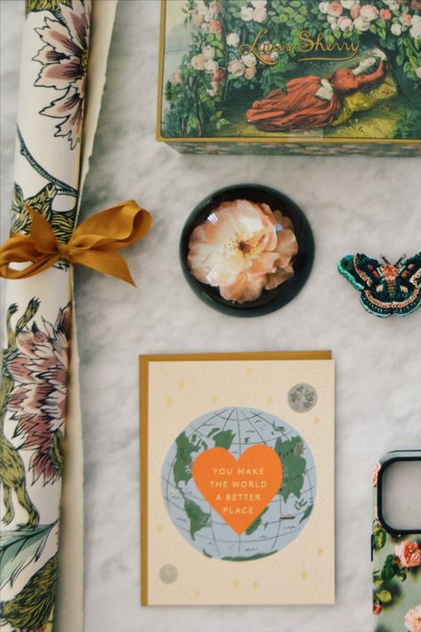 On a marble backdrop is a collection of items that give an earthy, romantic vibe. On the left, a floral art print is rolled up and tied with an amber-gold ribbon. At center is a blush-pink rose in bloom, framed in a crystal paperweight to look trapped in time. At the top, a woman in a red dress lies in a field of flowers on a decorative chocolate tin. To the right is a blue, pink, black beaded moth, and below is a rectangular card with a blue/green Earth on the middle, and a heart with text. Louis Sherry Tin, Museum Installation, Moth Brooch, Louis Sherry, Romantic Cottagecore, Paris Market, Jamie Beck, Cottagecore Gifts, Paris Markets