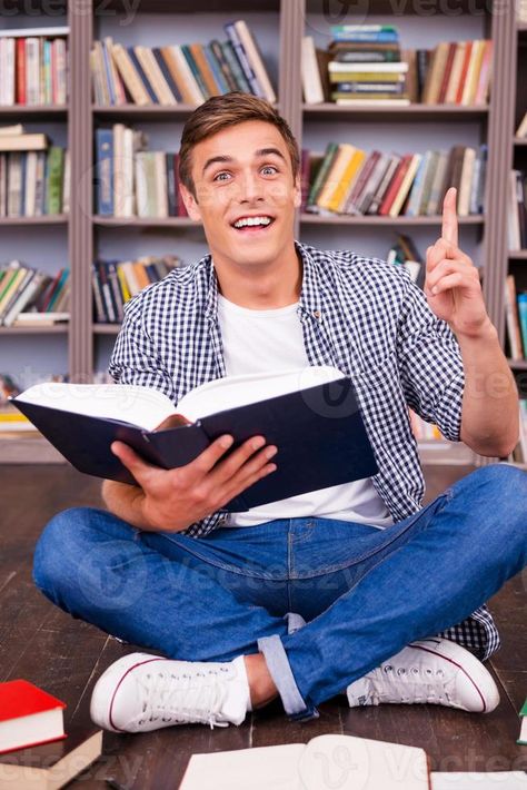 Bingo Happy young man holding book and gesturing while sitting against bookshelf Reading Book Refrence Pose, Person Holding Open Book, Book Holding Reference, Man Holding Book Pose Reference, Holding Open Book Reference, Person Holding Book Reference Drawing, Reading Book Pose Drawing Reference, Open Book Reference, Person Holding Book Reference