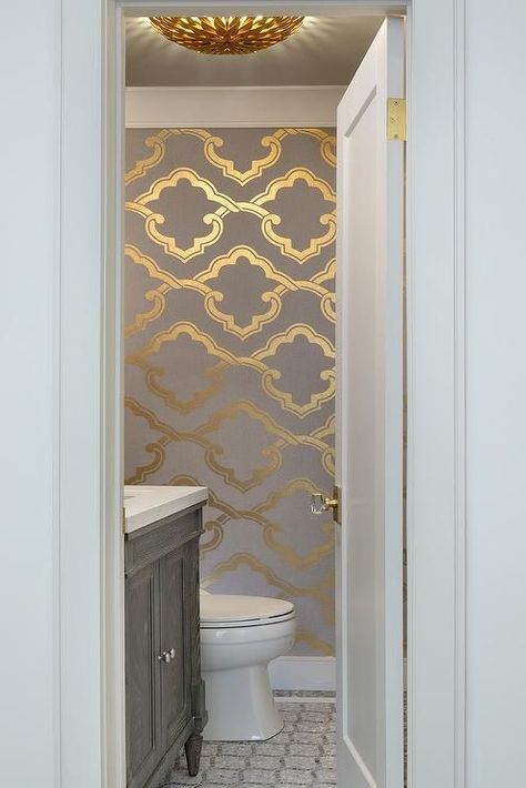 A white door finished with a glass door knob opens to a powder room boasting a gray mosaic tiled floor leading to a gray oak single washstand topped with white quartz surrounded by walls clad in gray and gold wallpaper. Light Gray Half Bathroom Ideas, Bathroom Gold Wallpaper, Gray And White Wallpaper Bathroom, Gold Wallpaper Powder Room, Gold Cloakroom, Gray Powder Room Ideas, White And Gold Powder Room, Mediterranean Powder Room Ideas, Gold Bathroom Wallpaper