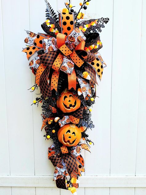 A large, stunning, whimsical Halloween swag wreath with Jack O Lanterns in the colors of yellow, orange, black and white will make a great addition to your décor this season!   Two pumpkins are featured in this dramatic swag wreath.  Created on a black teardrop swag, this wreath has lots of glitter and charm. Black glittery lacey ferns, black stems with glitter balls and candy corn can all be seen among the Halloween ribbons and streamers. A dramatic statement wherever you choose to display it! This large swag measures approximately 40" long, 18" wide and 8" deep. An original by Vine and Twine Studio and one of a kind, so if this is something you would like, don't hesitate as it can be yours. Halloween Swags, Halloween Swag, Lantern Wreath, Front Door Halloween, Halloween Theme Party, Holiday Swag, Decor Business, Teardrop Swag, Swag Wreath