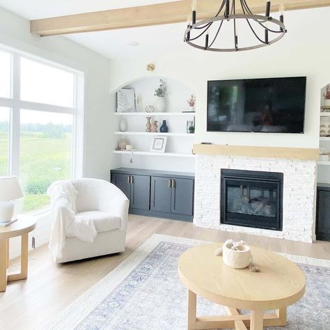 This custom-built living room wall features white arched shelving and black cabinets on a white fireplace wall. A mismatched white stone fireplace and blonde wood beam mantel give the room an airy feel, with a flatscreen television mounted above the mantel. White Fireplace Wall, Fireplace Wall Ideas With Tv, Arched Shelving, White Stone Fireplace, Fireplace Wall Ideas, White Stone Fireplaces, Beam Mantel, Built In Shelves Living Room, Condo Living Room