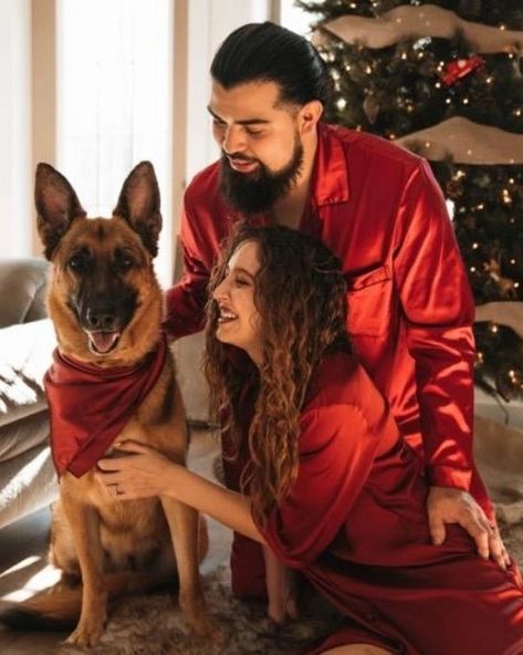 Christmas photoshoot. From left to right you'll see a beautiful German shepherd dog sitting to attention with red wine satin scarf with her human farming over her giving her attention and pats. Husband is a taller Hispanic man with longer black hair ties in the back with thick dark eyebrows and facial hair. He is wearing red wine satin long sleeve pajamas. His wife is a rather petite woman with voluminous curly hair smiling petting the pup smiling wearing red wine satin night shirt. X-Mas tree Couple Christmas Pictures With Dog Pajamas, Couples Christmas Photoshoot With Dog, Photoshoot For Christmas, Couple Christmas Photoshoot, Family Christmas Onesies, Couple Pajamas Christmas, Christmas Photoshoot Ideas, Dog Christmas Photos, Dog Christmas Pictures