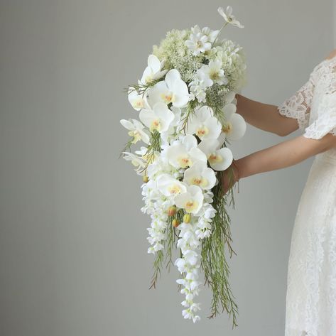 💐Product Description: This exquisite bridal bouquet showcases a stunning arrangement of white flowers, meticulously crafted to create a cascading effect. The bouquet features elegant white orchids as the focal point, complemented by smaller white blooms and delicate greenery. The design creates a waterfall-like appearance, adding a touch of sophistication and grace to any bridal ensemble. The flowers are thoughtfully arranged to ensure a balanced and harmonious look, with the cascading elements Wedding Flowers For Virgin Mary, White Flower Wedding Bouquet Dui, Silk Vs Real Flowers For Wedding, White Flowers And Greenery Bridal Bouquet, Calla Lily Bridal Bouquet, Orchid Bouquet Wedding, Teardrop Bouquet, Calla Lily Bridal, Cascading Bridal Bouquets