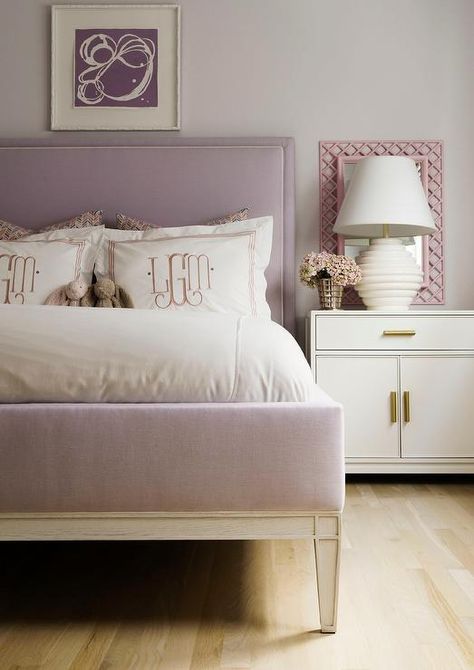 A purple abstract art piece hangs from a light gray wall over a lilac upholstered bed dressed in pink monogrammed bedding. Pink Purple Bedroom, Green Upholstered Bed, Pink And Purple Bedroom, White And Gold Nightstand, Lilac Bedding, Headboard With Lights, Purple Bedroom, Transitional Bedroom, Light Grey Walls