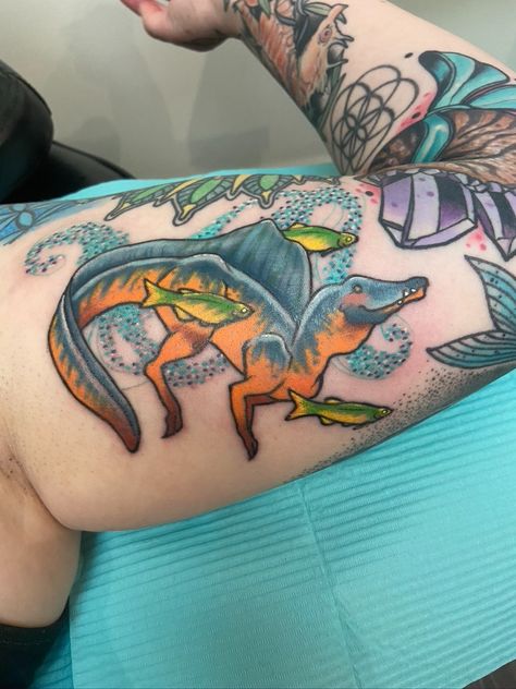 Tattoo is a spinosaurus swimming next to several small fish, background is glittery swirls in blues. Raptor Claw Tattoo, Carnotaurus Tattoo, Dinosaur Fossil Tattoo, Dilophosaurus Tattoo, Floral Dinosaur Tattoo, Traditional Dinosaur Tattoo, Ankylosaurus Tattoo, Spinosaurus Tattoo, Triceratops Tattoo