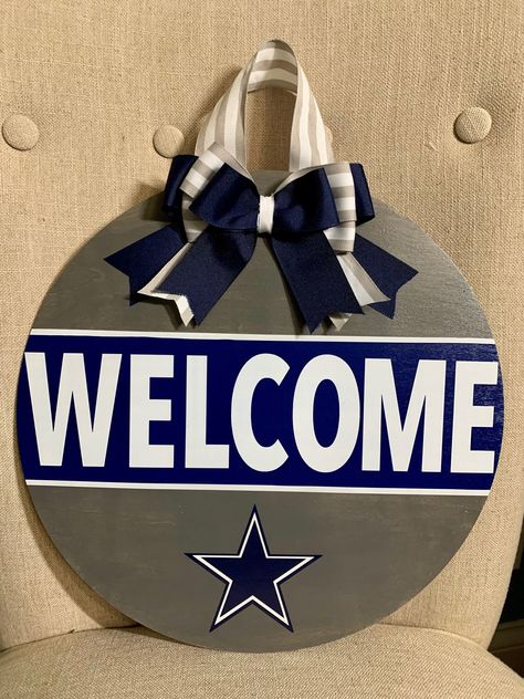 "Show your pride with this Cowboys Door Hanger! Just in time for the Playoffs!  Ready to hang, this is a 14\" wood round, hand painted with layered vinyl lettering and decor.  Coated with 2 layers of Polycrylic finish.  Ribbon and greenery may vary based on availability but will closely resemble the pictured.  If you would like to modify colors/decor/team name, feel free to message us!" Dallas Cowboys Round Wood Sign, Cowboys Door Hanger, Nfl Door Hanger, Dallas Cowboys Door Hanger, Dallas Cowboys Crafts, Tennessee Crafts, Dallas Cowboys Wreath, Cowboys Pictures, Cowboys Wreath