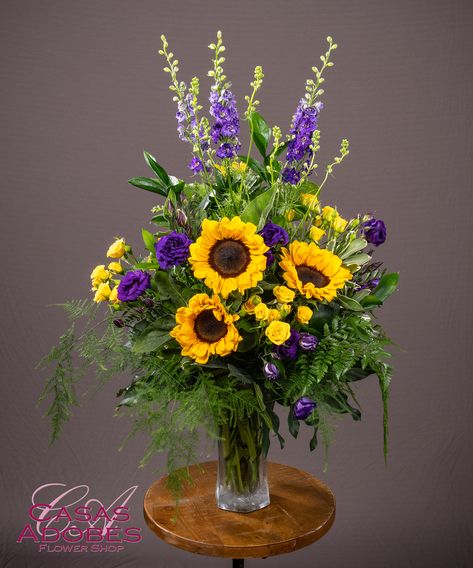 This Enduring Love Vase is designed with sunflowers, purple larkspur, purple lisianthus and yellow spray roses ina d beautiful vase. Sunflower Cemetery Arrangements, Sunflower And Purple Flower Arrangements, Purple And Gold Floral Arrangements, Fall Altar Flowers, Purple And Gold Flower Arrangements, Purple And Yellow Floral Arrangement, Floral Arrangements In Vases, Yellow And Purple Flower Arrangements, Sunflower Bouquet Vase